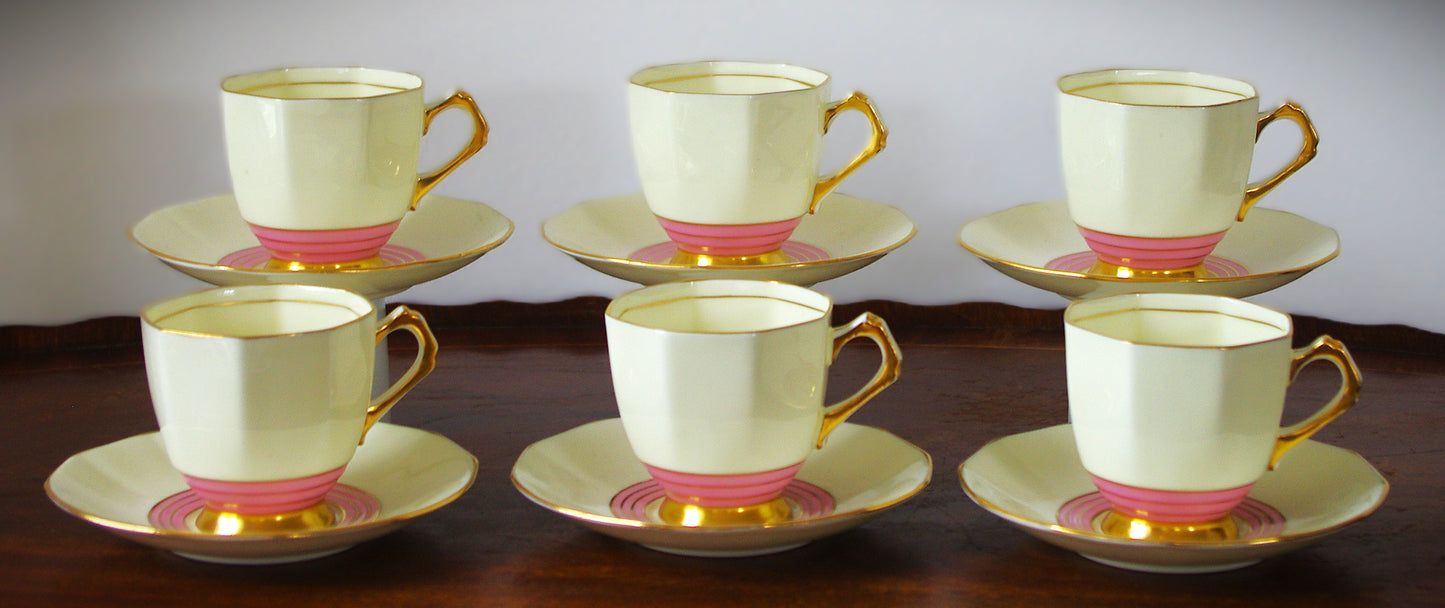 A set of six vintage Tuscan bone china tea cups and saucers from the 1920s, shown on a wooden tray. Each cup and saucer features an octagonal, faceted shape with a creamy ivory glaze, bold pink concentric bands at the base, and rich gold accents on the handles, rims, and bases. The close-up highlights the geometric gold handle and elegant Art Deco design. The maker’s mark on the underside reads “Plant Tuscan China Made in England 5788A.