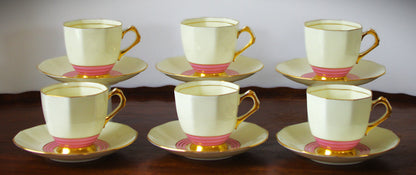 A set of six vintage Tuscan bone china tea cups and saucers from the 1920s, shown on a wooden tray. Each cup and saucer features an octagonal, faceted shape with a creamy ivory glaze, bold pink concentric bands at the base, and rich gold accents on the handles, rims, and bases. The close-up highlights the geometric gold handle and elegant Art Deco design. The maker’s mark on the underside reads “Plant Tuscan China Made in England 5788A.