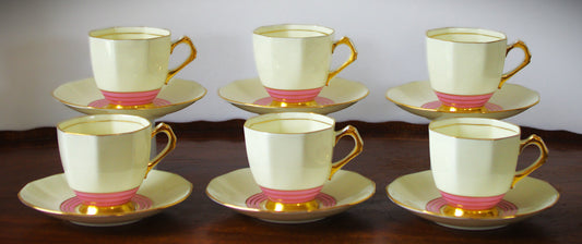 A set of six vintage Tuscan bone china tea cups and saucers from the 1920s, shown on a wooden tray. Each cup and saucer features an octagonal, faceted shape with a creamy ivory glaze, bold pink concentric bands at the base, and rich gold accents on the handles, rims, and bases. The close-up highlights the geometric gold handle and elegant Art Deco design. The maker’s mark on the underside reads “Plant Tuscan China Made in England 5788A.
